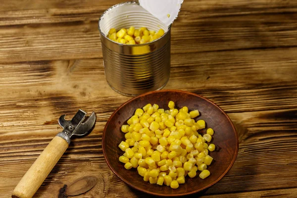 Open tin can and plate with corn and can opener on wooden table — Stock Photo, Image