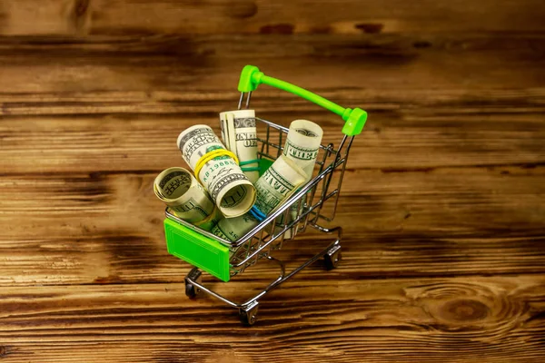 Pequeño carrito de compras con rollos de billetes de cien dólares sobre fondo de madera. Concepto de consumo —  Fotos de Stock