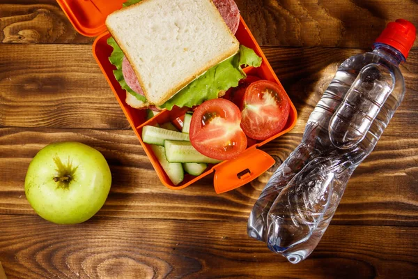 学校午餐盒，木桌上摆着三明治和新鲜蔬菜、一瓶水和青苹果。健康饮食理念。顶部视图 — 图库照片
