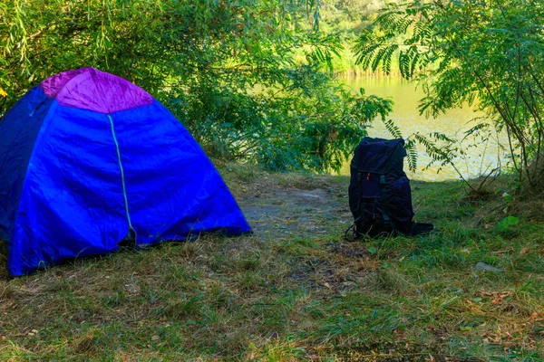 Mavi kamp çadırı ve sırt çantası bir nehir yakınında ormanda — Stok fotoğraf
