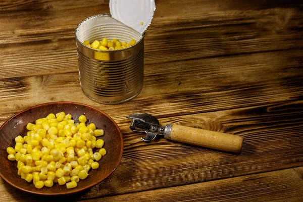 Open tin can and plate with corn and can opener on wooden table — Stock Photo, Image