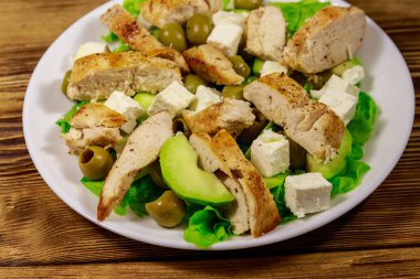 Ahşap masada kızarmış tavuk göğsü, yeşil zeytin, beyaz peynir, avokado, marul ve zeytinyağı ile lezzetli salata