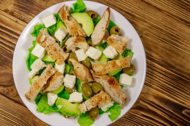 Lezzetli salata, kızarmış tavuk göğsü, yeşil zeytin, feta peyniri, avokado, marul ve ahşap masada zeytinyağı. Üst görünüm