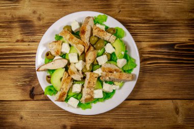 Lezzetli salata, kızarmış tavuk göğsü, yeşil zeytin, feta peyniri, avokado, marul ve ahşap masada zeytinyağı. Üst görünüm
