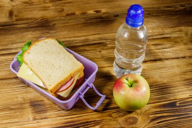 Sandviçli beslenme çantası, bir şişe su ve ahşap masada elma.