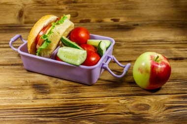 Elma, beslenme çantası, hamburgerler ve ahşap bir masada taze sebzeler.