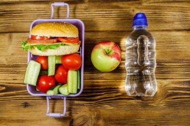 Bir şişe su, elma ve beslenme çantası, içinde hamburgerler ve ahşap bir masada taze sebzeler. Üst görünüm