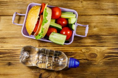 Bir şişe su ve hamburgerli beslenme çantası ve ahşap bir masada taze sebzeler. Üst görünüm