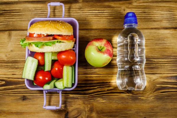 Bir şişe su, elma ve beslenme çantası, içinde hamburgerler ve ahşap bir masada taze sebzeler. Üst görünüm