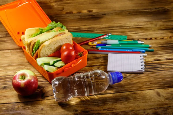 Back to school concept. School supplies, bottle of water, apple and lunch box with sandwiches and fresh vegetables on a wooden desk