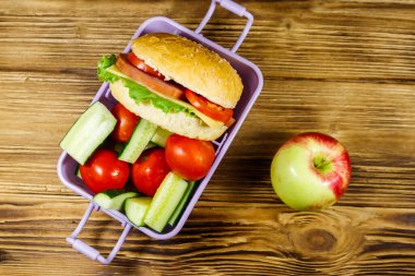 Elma, hamburger ve ahşap masada taze sebzeli beslenme çantası. Üst görünüm