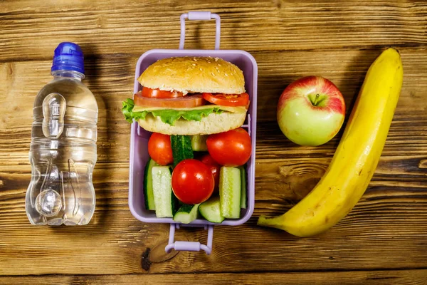 Bir şişe su, elma, muz ve hamburgerli beslenme çantası ve tahta bir masada taze sebzeler. Üst görünüm