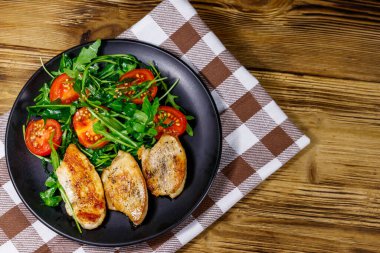 Kızarmış tavuk göğsü, taze roka salatası ve ahşap masada vişneli domates. Üst görünüm