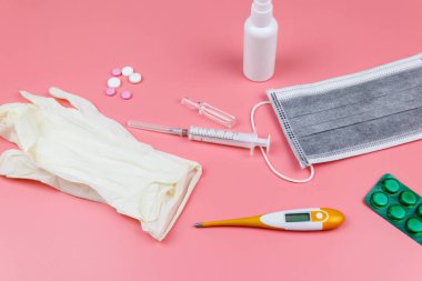 Koronavirüsü önleme ve tedavi etme kavramı. Tıbbi maske, el dezenfektanı, elektronik termometre, haplar, şırınga, ampuller, pembe arka planda tıbbi eldivenler.