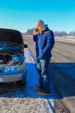 Kışlık yolda bozuk bir arabanın yanında duran üzgün genç adam araba servisini arıyor.