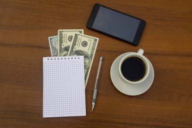 Bir fincan kahve, akıllı telefon, dolar, not defteri ve tahta masadaki kalem. Mali planlama kavramı