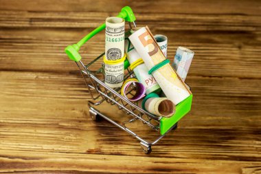 Shopping cart with money from different countries: euros, us dollars, ukrainian hryvnia, russian rubles, egyptian pounds on wooden background clipart