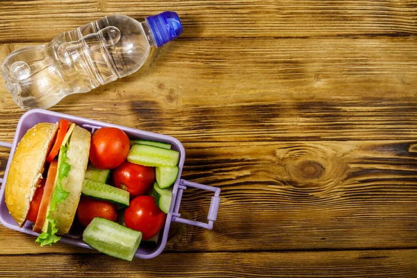 Bottle of water and lunch box with burgers and fresh vegetables on a wooden table. Top view, copy space