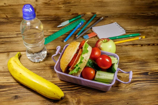 Back to school concept. School supplies, bottle of water, apple, banana and lunch box with burgers and fresh vegetables on a wooden table