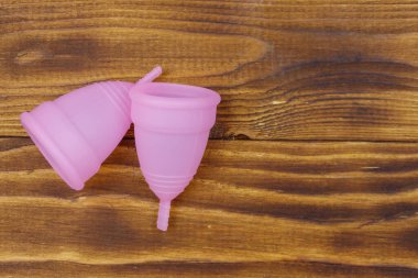Ahşap arka planda tekrar kullanılabilen pembe silikon adet bardakları. Üst görünüm, boşluğu kopyala. Kadın hijyeni, jinekoloji ve sağlık kavramı