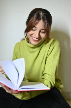 Kitap okuyan güzel genç bir kadın. Gülümseyen kadın kitap okuyor. Kitap okumak ilginçtir. Kadehler ellerde. Beyaz duvar. Okuma zamanı. Kitapla eğlen.