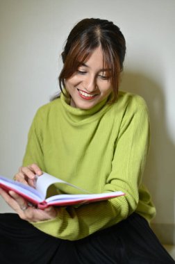 Kitap okuyan güzel genç bir kadın. Gülümseyen kadın kitap okuyor. Kitap okuyorum. Kadehler ellerde. Beyaz duvar. Okuma zamanı. Kitapla eğlen.