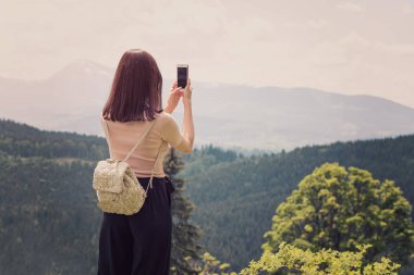 Dağ ve orman bir smartphone fotoğraf üzerinde bir sırt çantası ile kız. Arkadan görünüm. Güneşli yaz gün