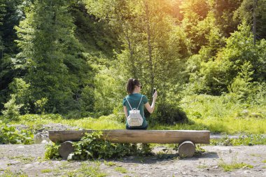 Kız ormanın yakınındaki bir sırt çantası ile bir smartphone bir fotoğrafı çeker. Arkadan görünüm. Yaz güneşli gün