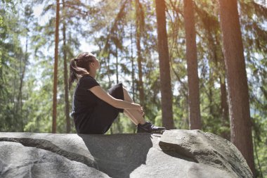 Kız ormanın içinde büyük bir kayanın üzerinde oturuyor. Yaz güneşli gün. Alt görünümü