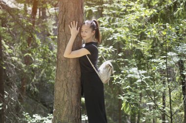 Bir sırt çantası ile kız ormanda kapalı gözleri ile ağaca sarılmak. Yaz güneşli gün.