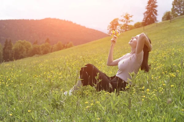 芝生の上に座っている野生の花の花束とかなり若い女性 背景に山々 夏の晴れた日 — ストック写真