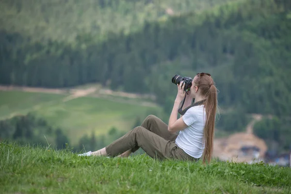 Girl with camera sits on a hill and photography nature. Summer day