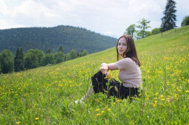Genç ve güzel kadınla çimenlerin üzerine oturmuş kır çiçekleri buketi. Arka planda dağlar. Yaz güneşli gün