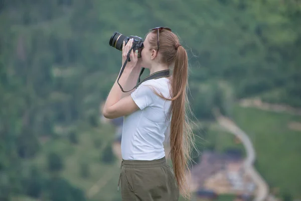 Girl with camera stands on a hill and photographing nature. Forest on background