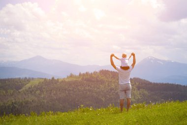 Onun omuzlarında küçük oğlu olan babası. Arka planda dağlar. Yaz güneşli gün. Arkadan görünüm