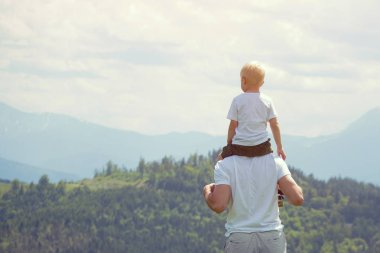 Onun omuzlarında küçük oğlu olan babası. Arka planda dağlar. Yaz güneşli gün. Arkadan görünüm
