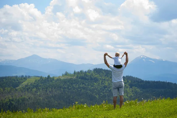 Onun omuzlarında küçük oğlu olan babası. Arka planda dağlar. Yaz güneşli gün. Arkadan görünüm