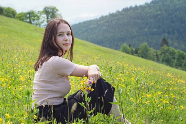 Genç ve güzel kadınla çimenlerin üzerine oturmuş kır çiçekleri buketi. Arka planda dağlar. Yaz güneşli gün