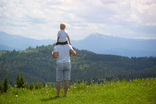 Onun omuzlarında küçük oğlu olan babası. Arka planda dağlar. Yaz güneşli gün. Arkadan görünüm