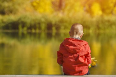 İskelede oturup elinde yaprakları kırmızı ceketli çocuk. Sonbahar, güneşli. Arkadan görünüm