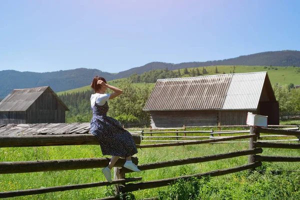 Young woman sitting on a wooden fence. Houses and green mountains in the background