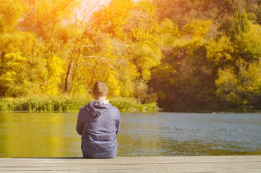 Genç adam iskelede oturur. Sonbahar güneşli gün. Arkadan görünüm