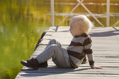 Sarışın küçük oğlan iskelede oturuyor. Sonbahar güneşli gün. Nehir banka