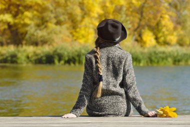 İskelede oturan şapkalı kız. Sonbahar günü. Arkadan görünüm