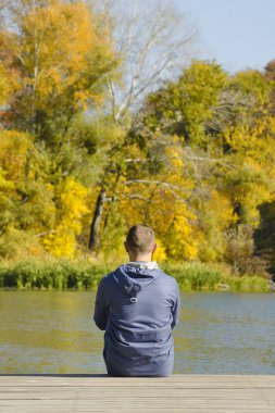 Genç adam iskelede oturur. Sonbahar güneşli gün. Arkadan görünüm