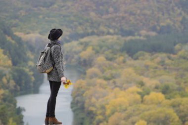 Bir sırt çantası ve bir tepe üzerinde bir şapka duran kız. Ellerini kaldırdı. Nehir ve dağ aşağıda. Yan görünüm