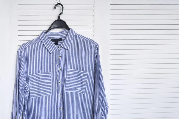 Bllue striped shirt hanging on a hanger. White wooden screen on the background. Fashionable wardrobe