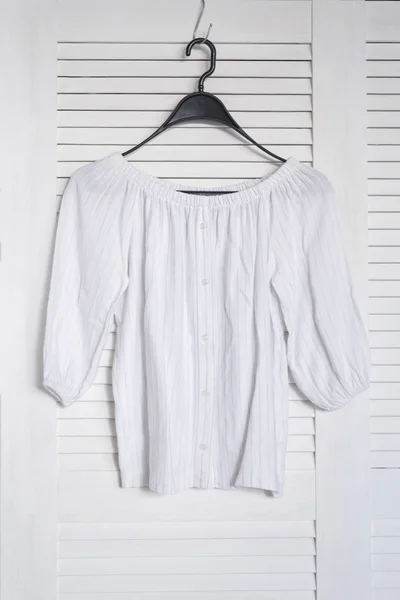 White blouse hanging on a hanger. White wooden screen on the background. Fashionable wardrobe