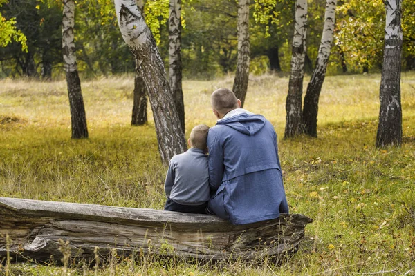 Father and son are sitting in the autumn forest on a log. Back view