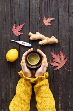 Bir fincan çay eller içinde. Limon, zencefil ve termometre. Soğuk tedavi. Ahşap arka plan. Üstten görünüm